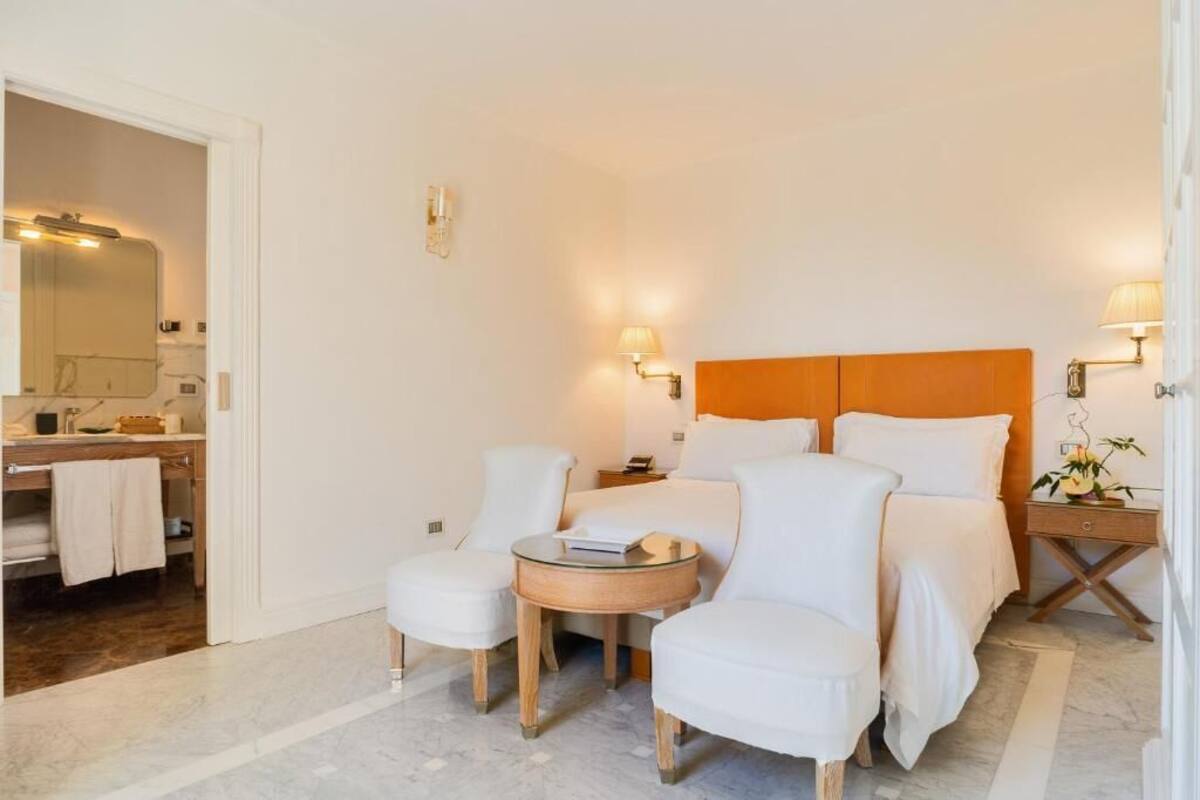 A hotel room with a bed, two white armchairs, a small round table, a nightstand, and a bathroom visible through an open door.