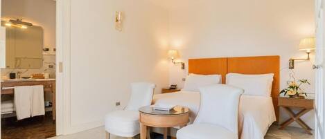 A hotel room with a bed, two white armchairs, a small round table, a nightstand, and a bathroom visible through an open door.