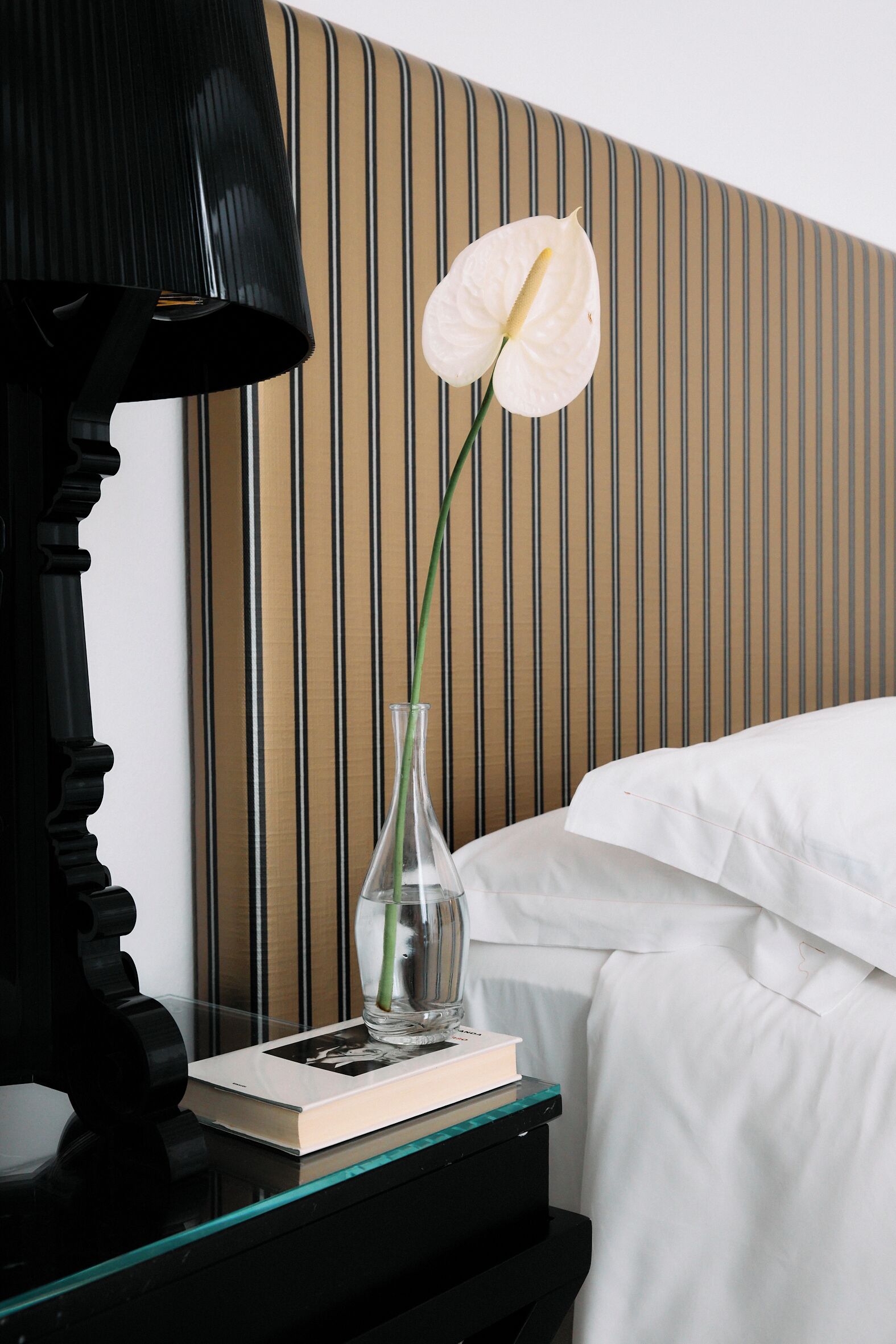 A neatly made bed with white linens, a black lamp on a bedside table, a clear vase with a single white flower, and a book.