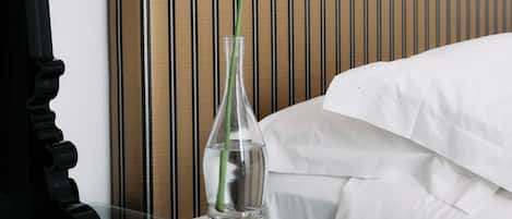 En pænt redt seng med hvide sengetæpper, en sort lampe på et natbord, en klar vase med en enkelt hvid blomst og en bog.