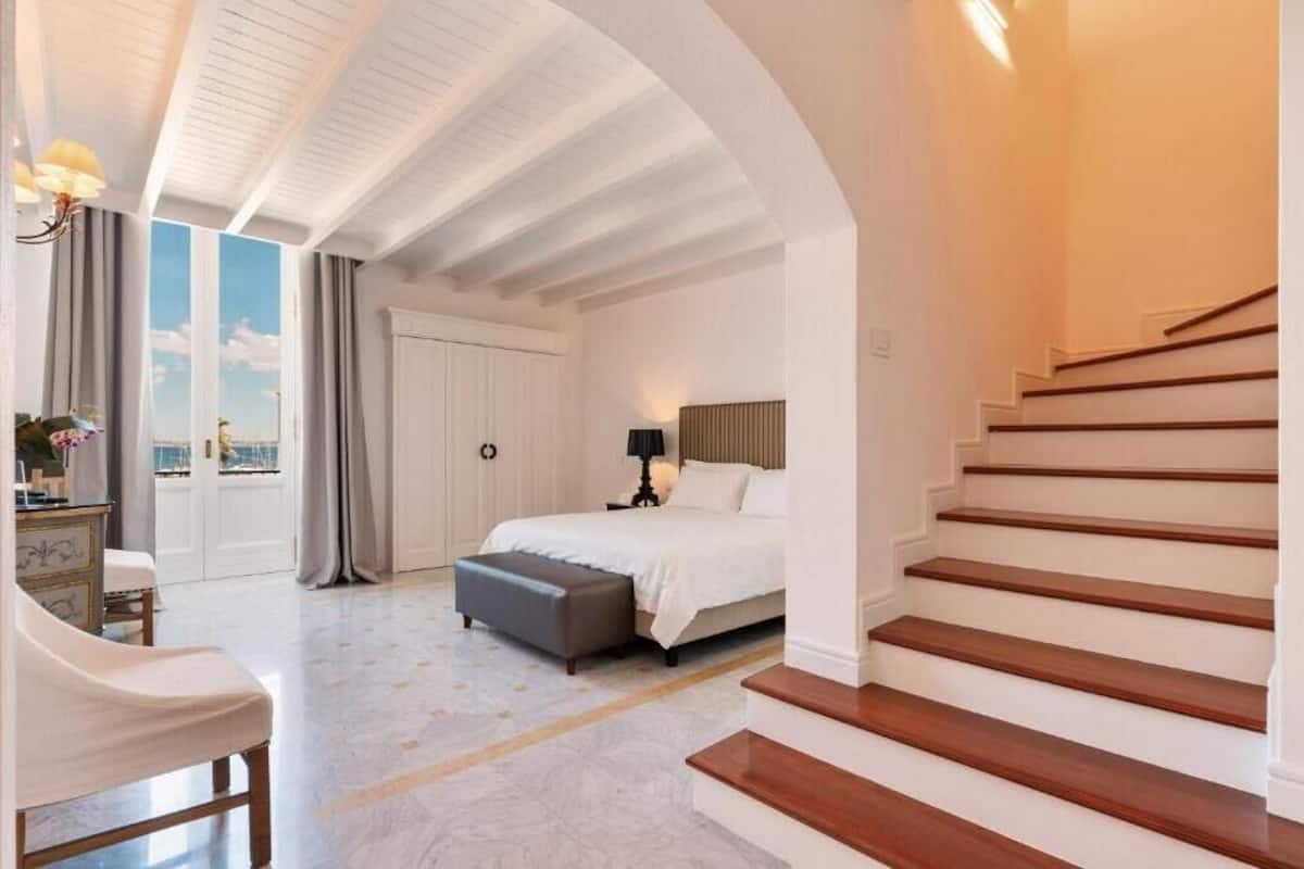 A bedroom with a bed, a nightstand, a lamp, a window with curtains, a staircase, and a view of the sky.