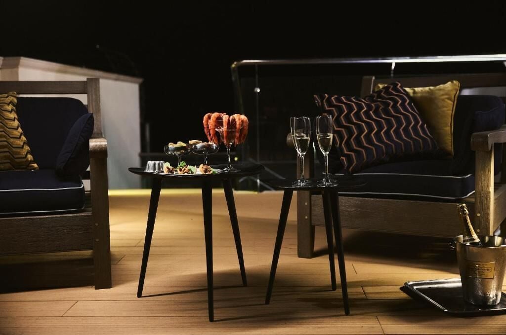 Uma sala de estar com decoração escura, um sofá, uma mesinha com comida e bebidas, e um objeto decorativo que se assemelha a um caranguejo.