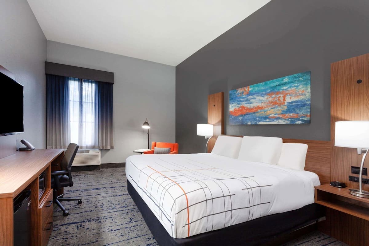 Ein Hotelzimmer mit Bett, Schreibtisch, Stuhl, Lampe und Kunstwerken.