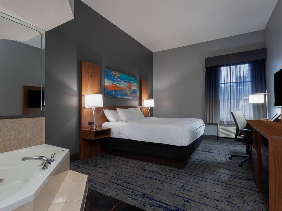 A hotel room with a large bed, two bedside lamps, a desk with a chair, a TV, and a bathroom with a bathtub and sink.
