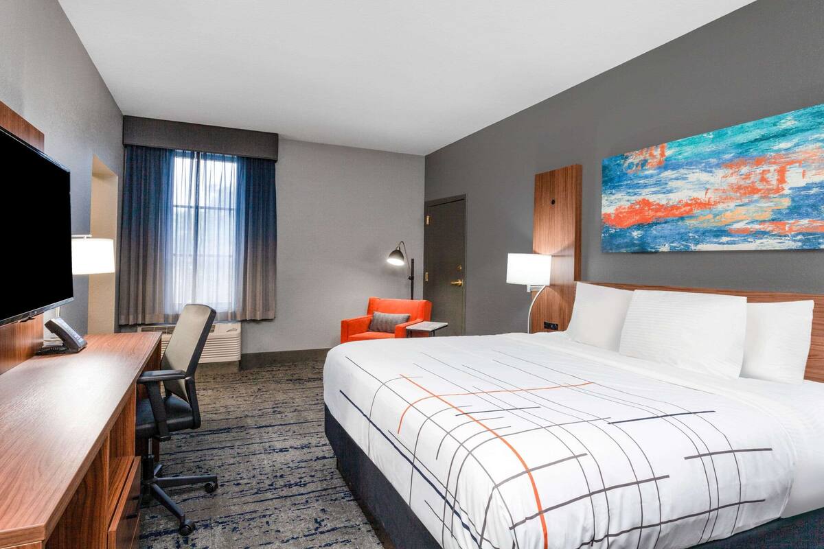 Ein Hotelzimmer mit einem großen Bett, einem Schreibtisch mit Computer, einem Sessel, einem Fernseher und einem Bild an der Wand.