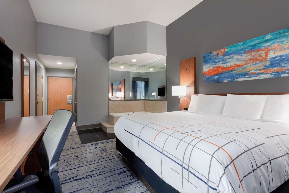 Ein Hotelzimmer mit Bett, Schreibtisch, Stuhl, Fernseher, einem Gemälde und einem durch eine Glasscheibe sichtbaren Badezimmer.