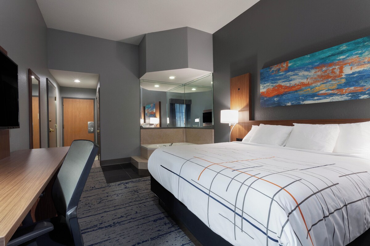 A hotel room with a large bed, a desk, a chair, a mirror, and a painting on the wall.