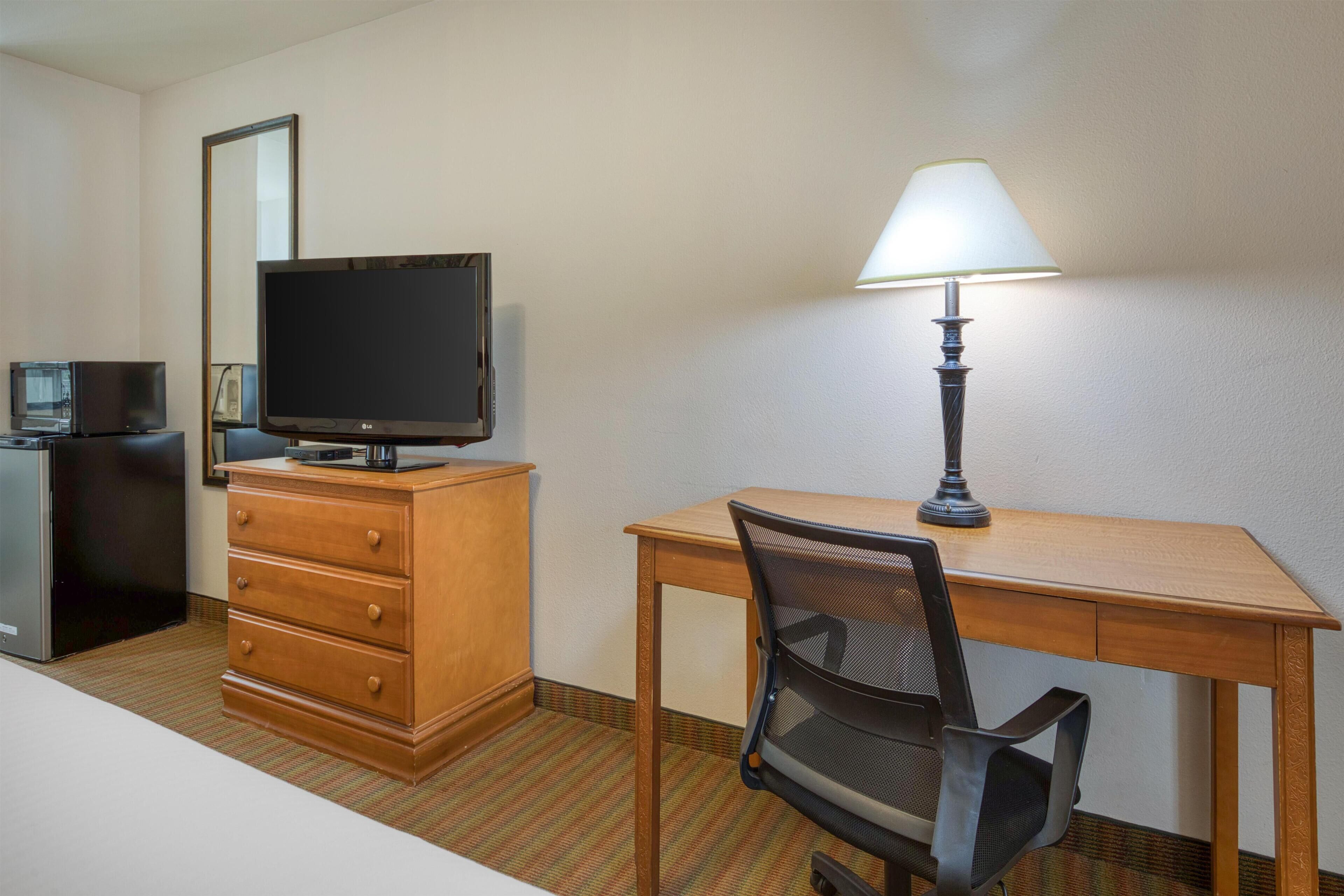 Habitación de hotel con escritorio de madera, una silla, televisor, lámpara, cómoda y microondas.