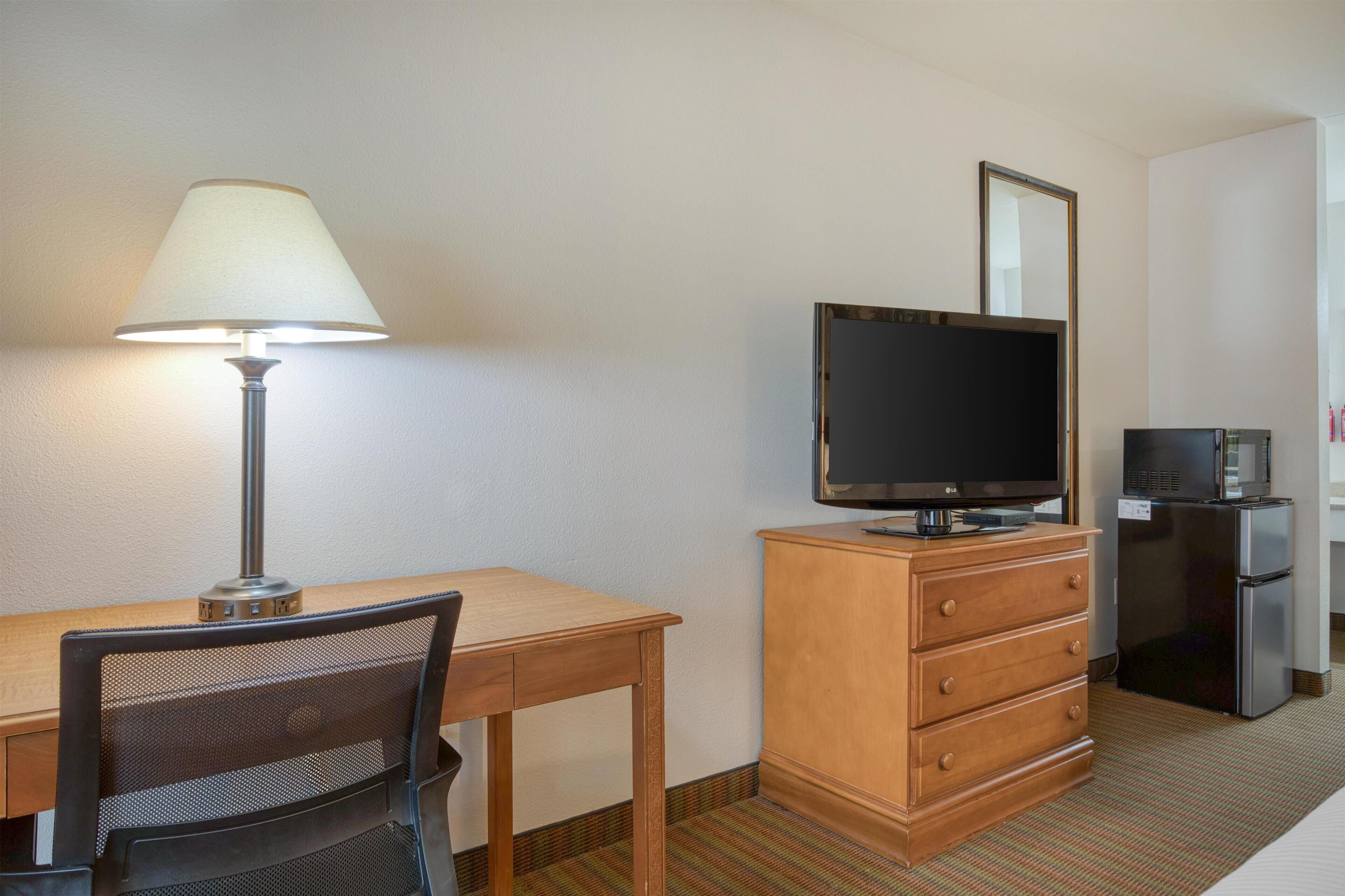 A hotel room with a wooden desk, a chair, a television on a wooden cabinet, and a refrigerator.
