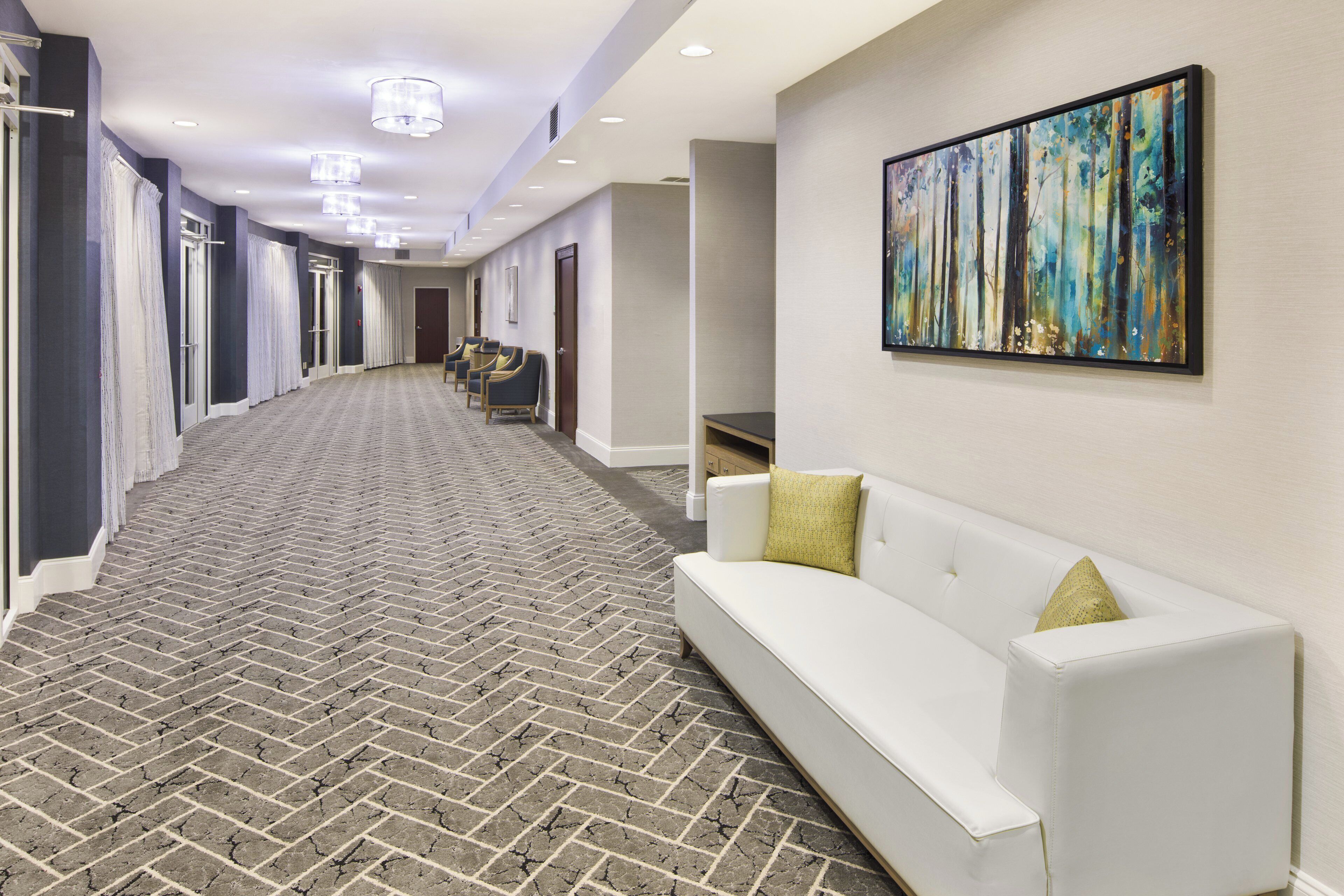 A hotel corridor with a patterned carpet, a white sofa with cushions, and a painting on the wall.