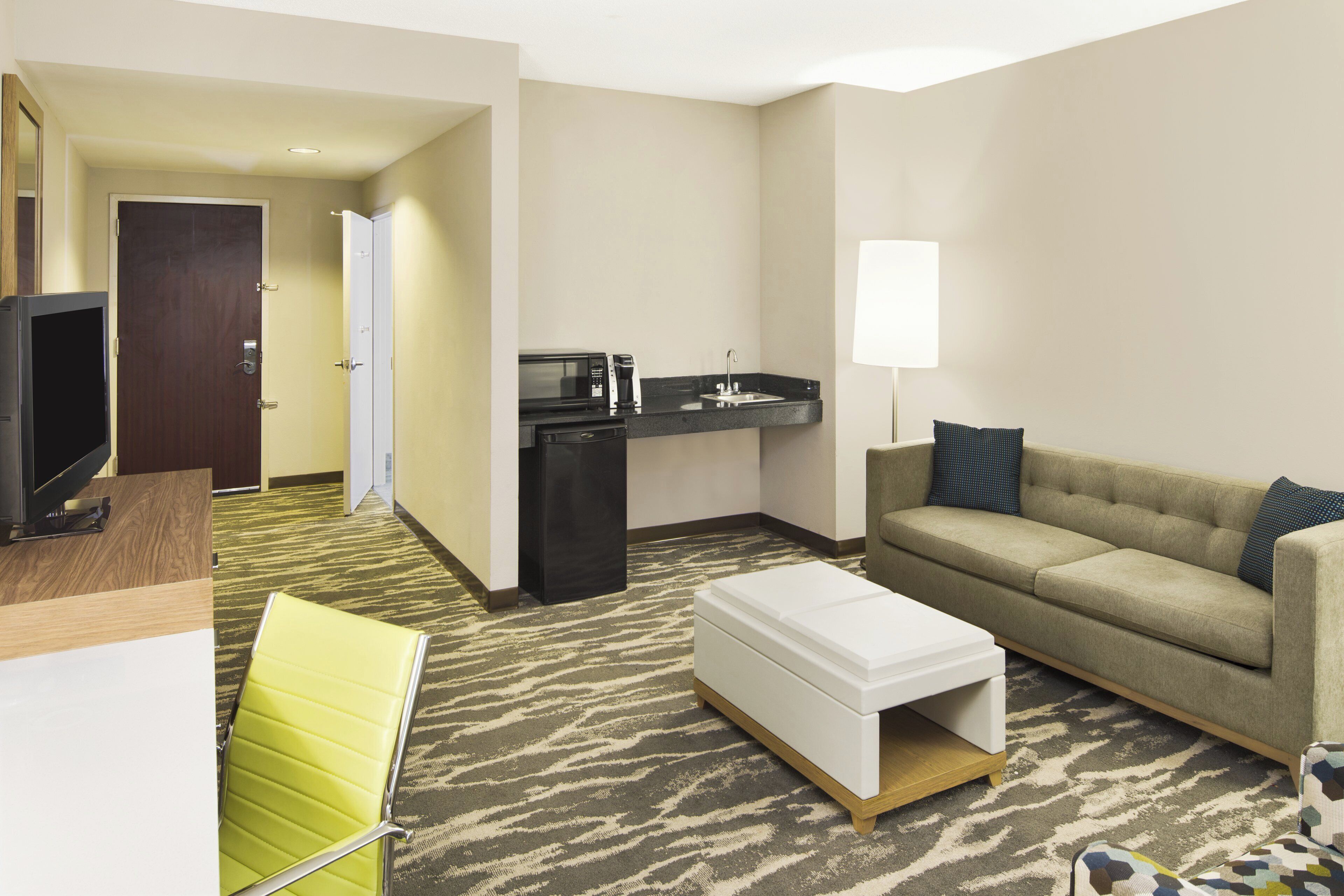 A hotel room with a kitchenette, a sofa, a TV, and a patterned carpet.