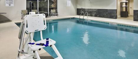 Una piscina interior con un elevador de piscina, un cartel de prohibición de clavados y un letrero de precaución en la pared.