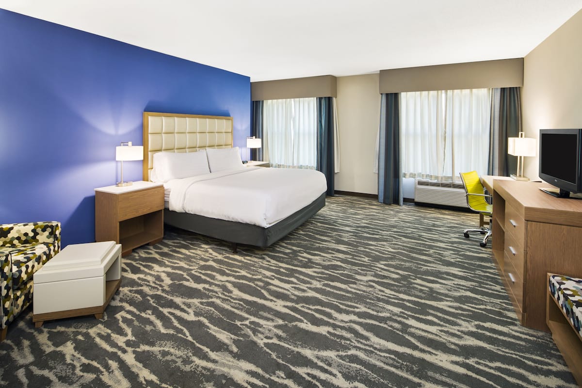 A hotel room with a large bed, a desk with a computer, a chair, a TV, and a window with curtains.