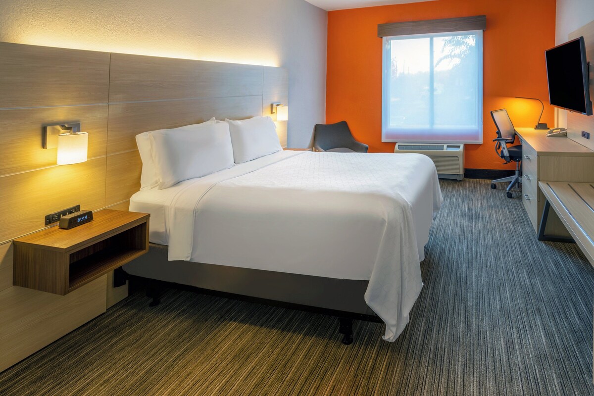 A hotel room with a bed, bedside table, lamp, TV, desk, chair, and window.