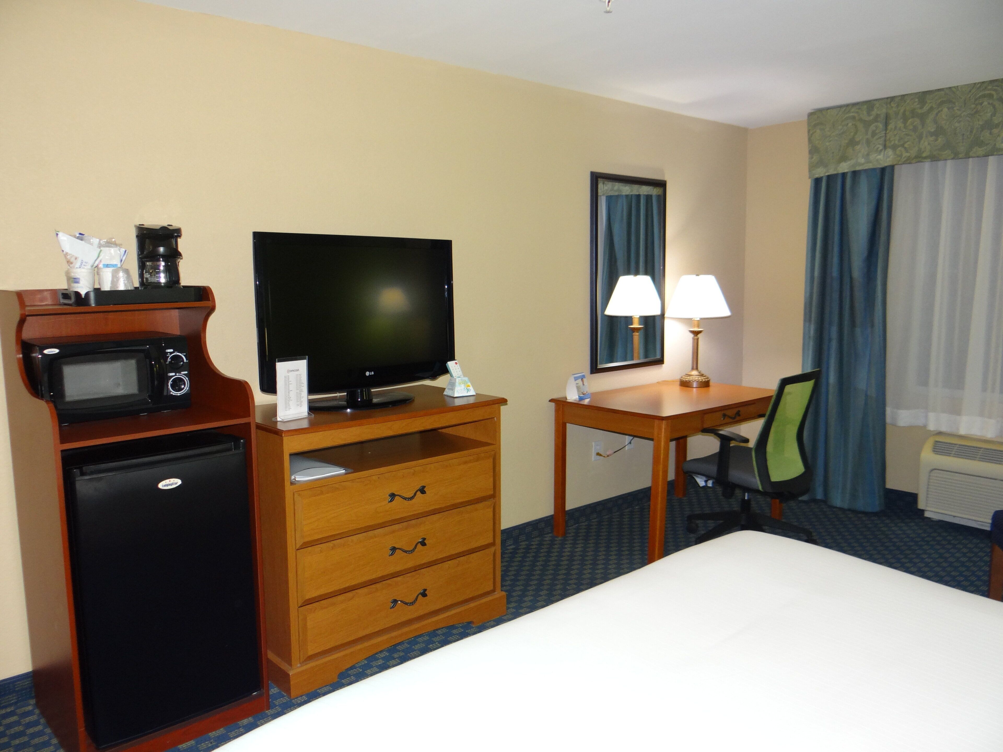 Ein Hotelzimmer mit Bett, Schreibtisch, Fernseher, Mikrowelle, Kühlschrank, Lampe und Stuhl.