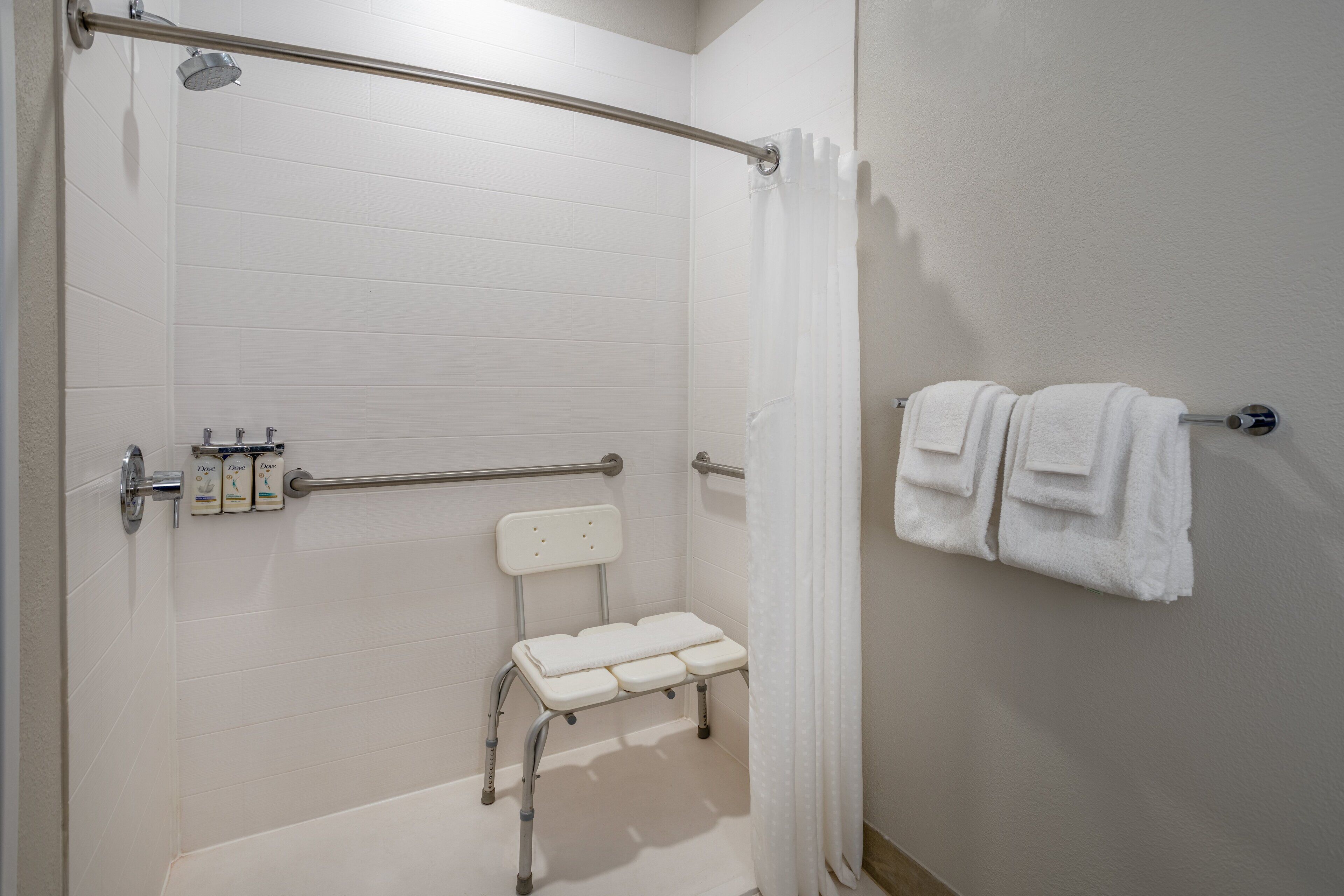 Un baño con ducha blanca, barra de apoyo, banco blanco y toallero con toallas.