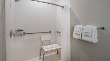 Un baño con ducha blanca, barra de apoyo, banco blanco y toallero con toallas.