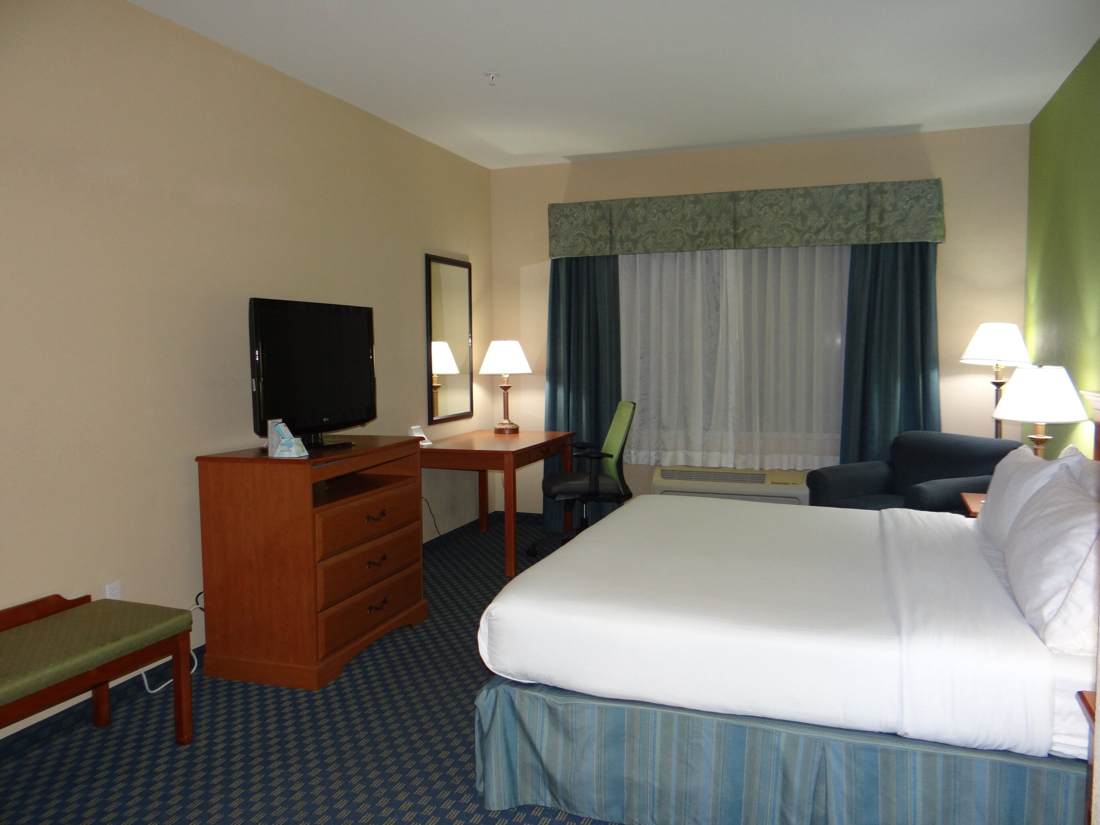 A hotel room with a bed, a television, a desk, two lamps, a chair, and a window with curtains.