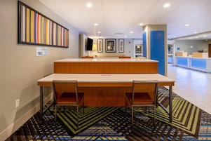 A hotel reception area with a reception desk, chairs, a patterned carpet, and a wall with a vertical art piece.