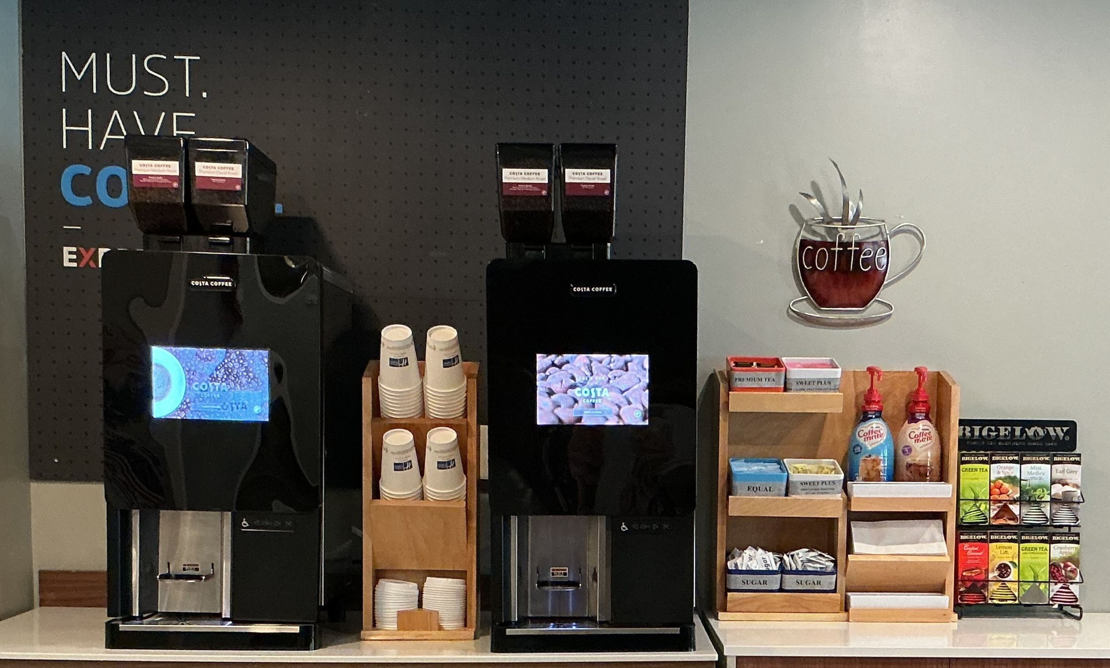 Dos máquinas de café, un expositor de vasos desechables y una estantería con productos para el café.