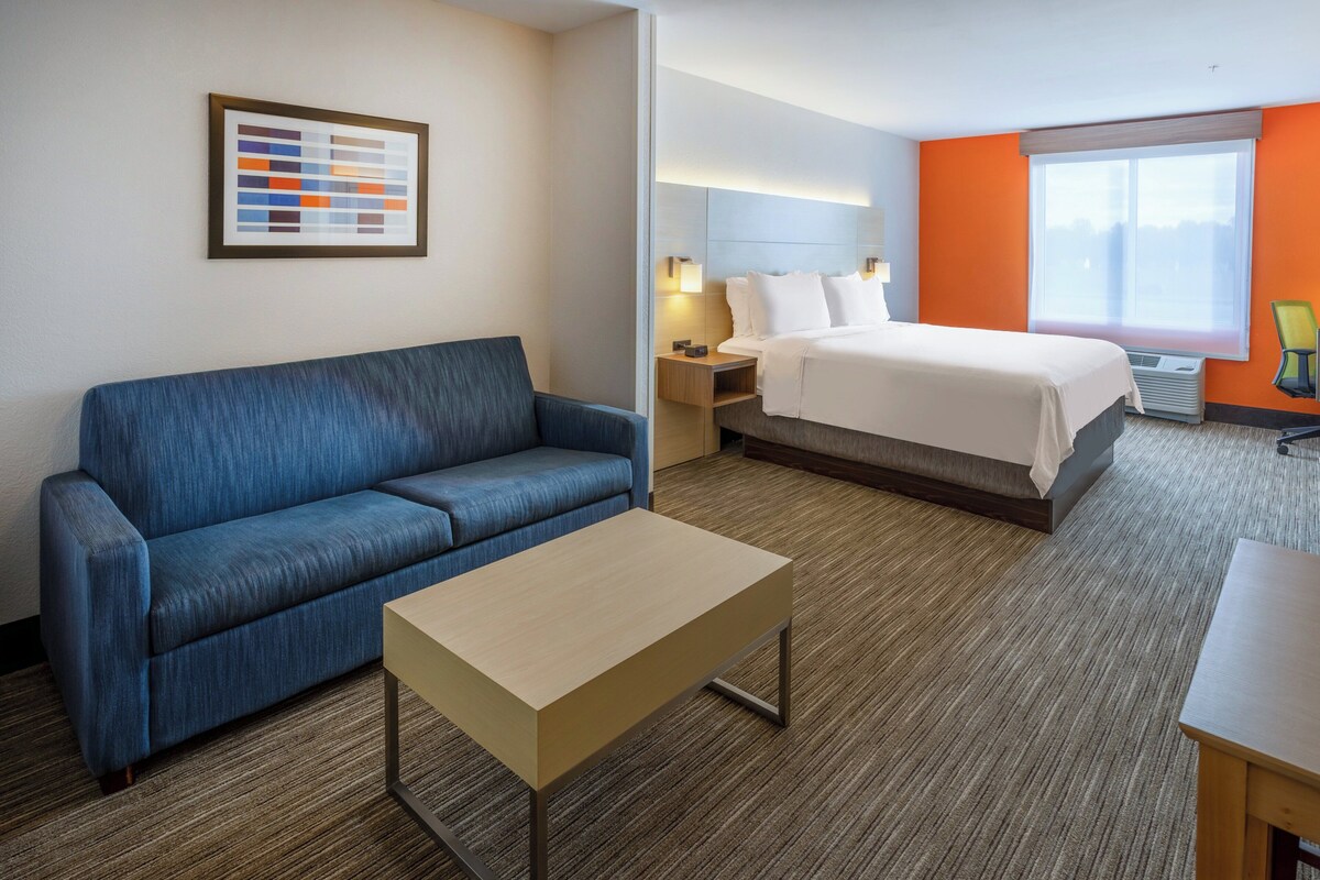 A hotel room with a blue sofa, a small wooden table, a bed with white linens, and a large window with orange walls.