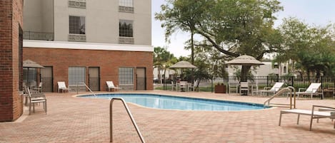 A swimming pool with a brick patio, lounge chairs, and a building with a brick facade and large windows.