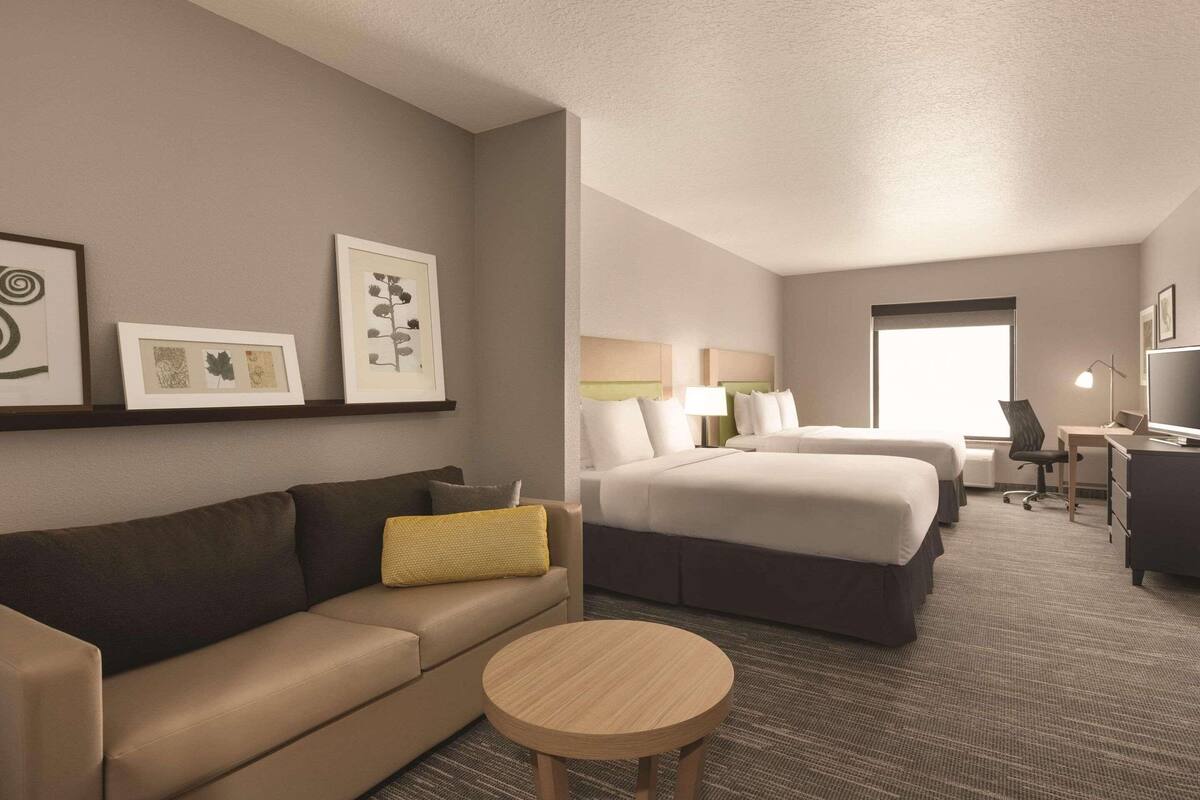 A hotel room with a sofa, a round table, a bed with two pillows, a desk with a chair, and framed artwork on the wall.