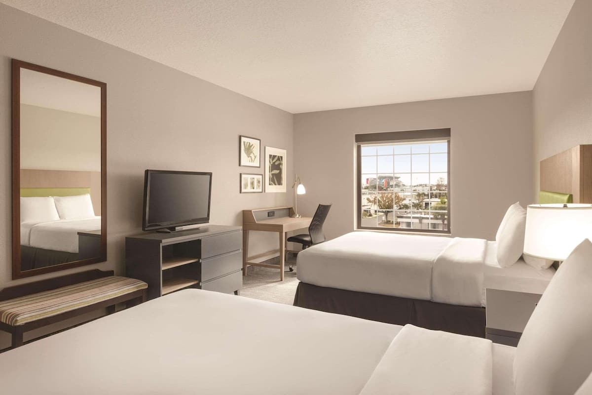 A hotel room with two beds, a desk with a chair, a television, and a large window offering a city view.