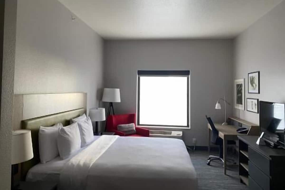 A hotel room with a bed, a desk, a chair, a lamp, and a window.
