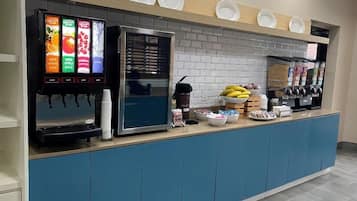 A self-service beverage station with multiple drink dispensers, a refrigerator, and a variety of food items on the counter.