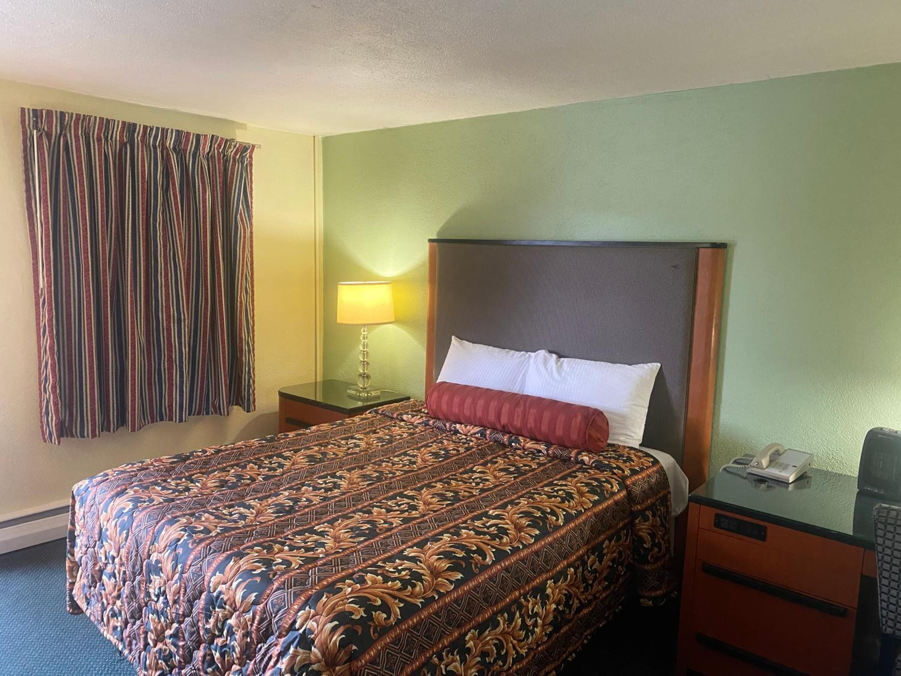 Ein Hotelzimmer mit einem großen Bett, einem Nachttisch mit Lampe, einem Telefon und einem Schreibtisch mit Computer.