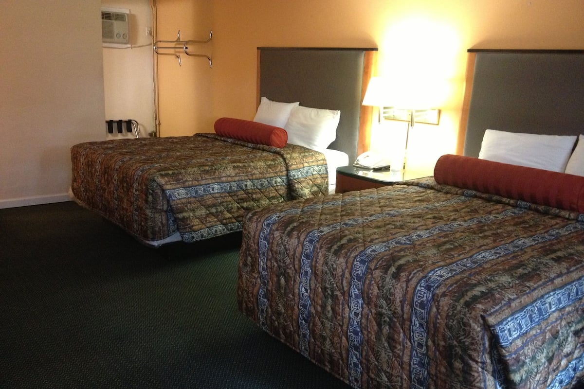 Two double beds with patterned bedding, a bedside table with a lamp, and a wall-mounted air conditioning unit.