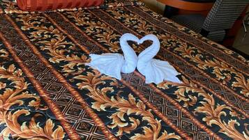 A neatly made bed with a decorative bedspread, two towels arranged in a heart shape, a red pillow, and a small bedside table with a lamp and a phone.