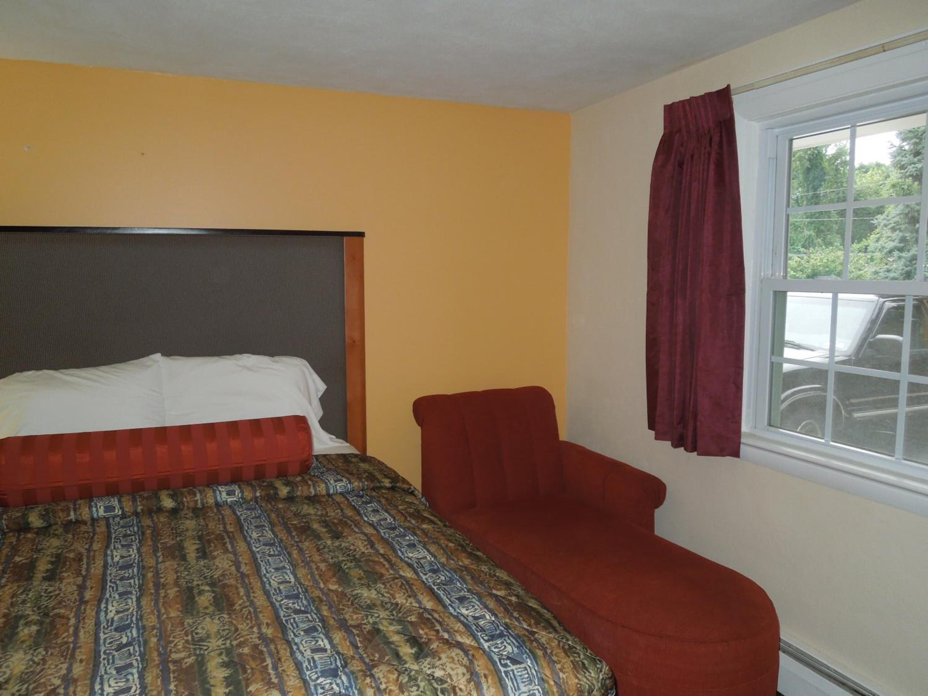 A room with a bed, a red chair, and a window with a view of trees and a car.