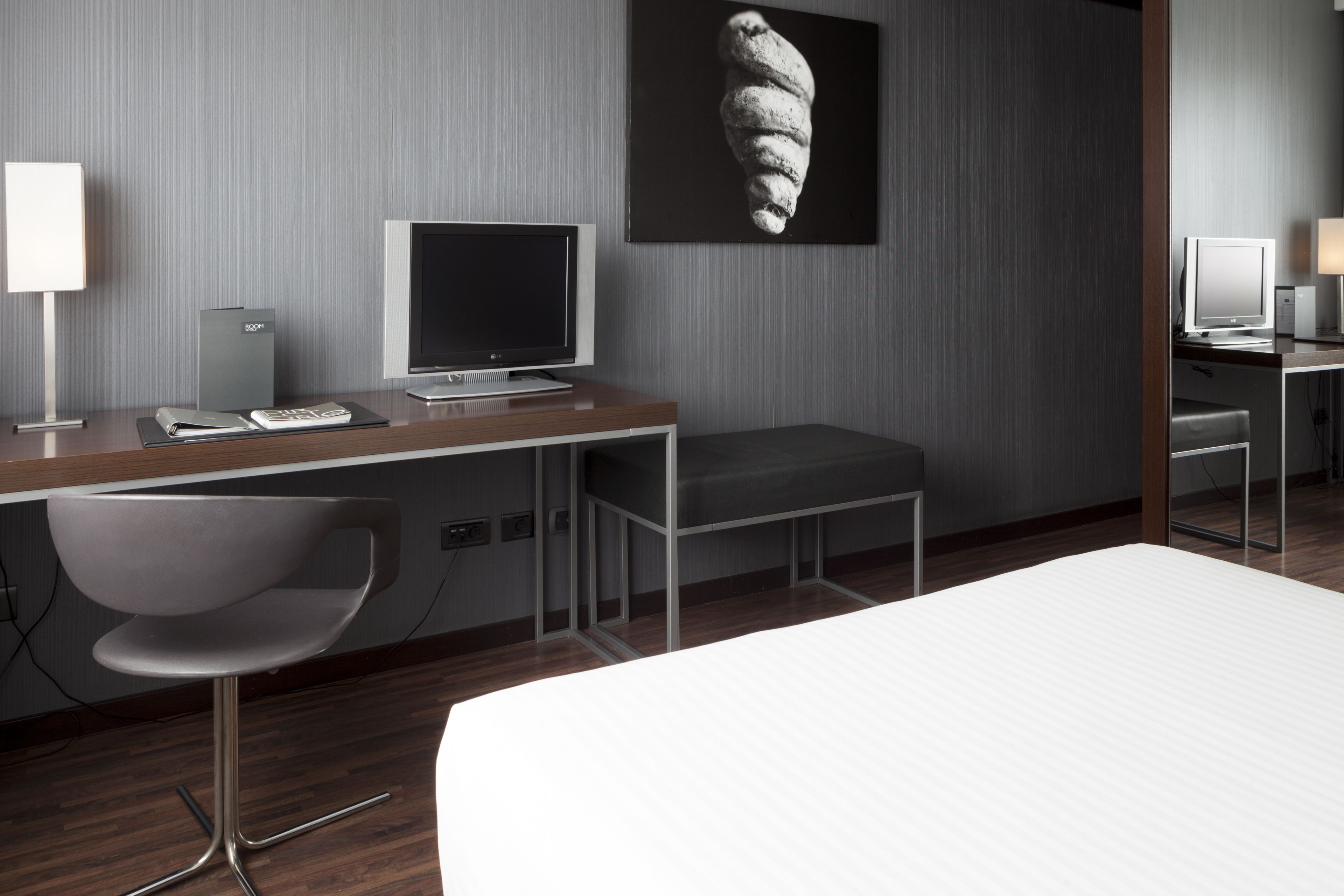 Une chambre d’hôtel moderne équipée d’un bureau en bois, d’un ordinateur portable, d’un téléviseur à écran plat, d’un fauteuil confortable, d’un lit et d’une grande œuvre d’art accrochée au mur.