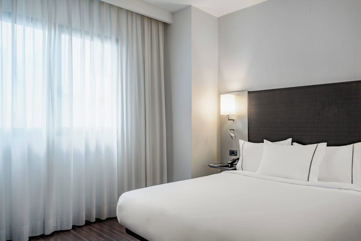 A hotel room with a large bed, a nightstand, a lamp, and sheer curtains.