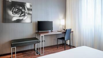 A hotel room with a bed, a desk with a flat-screen TV, a chair, a bench, and a large abstract painting on the wall.