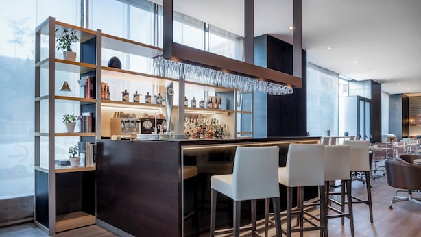 Uma área de bar moderna com balcão de madeira escura, banquetas brancas e prateleiras com livros e garrafas.