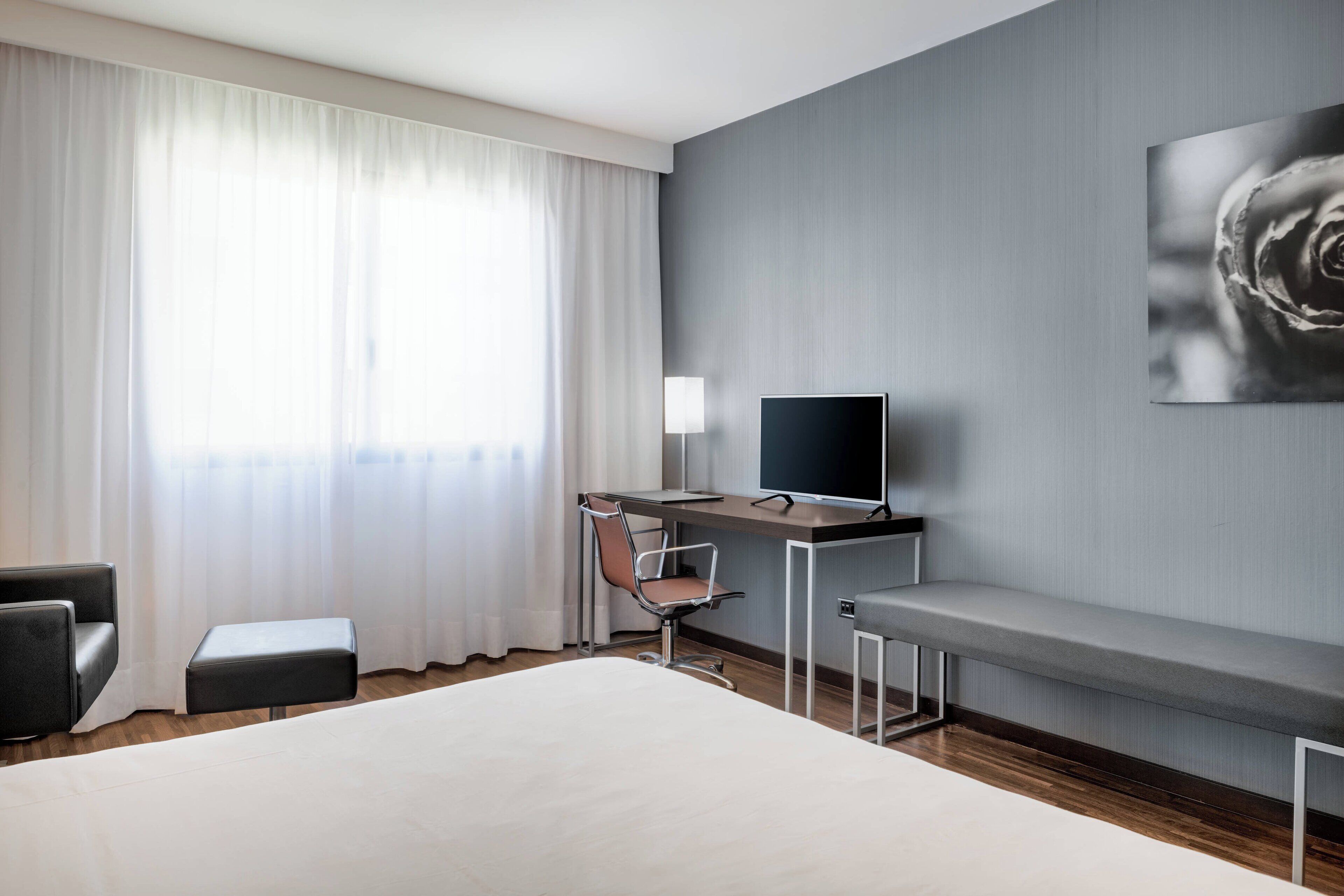 A modern hotel room with a large bed, a desk with a computer, a chair, and a bench.