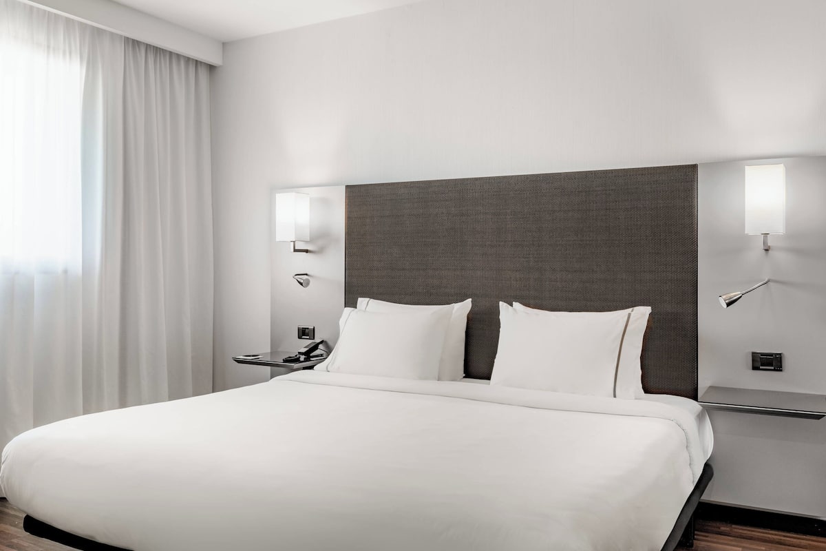 A modern hotel room with a large bed, a nightstand, a lamp, and a view of the city through the window.