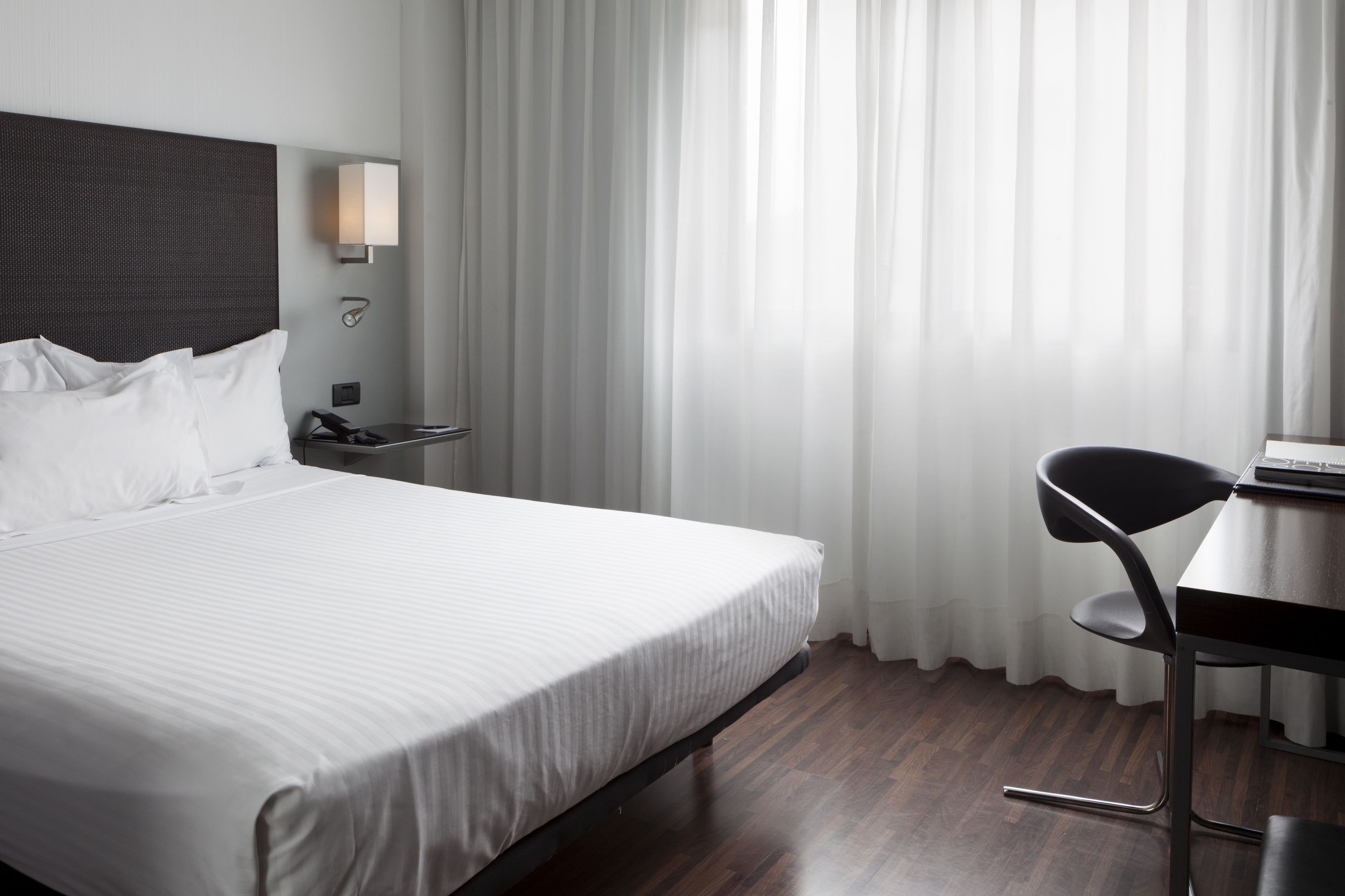 Habitación de hotel con una cama grande, una mesita de noche con lámpara, una silla y un escritorio con computadora.