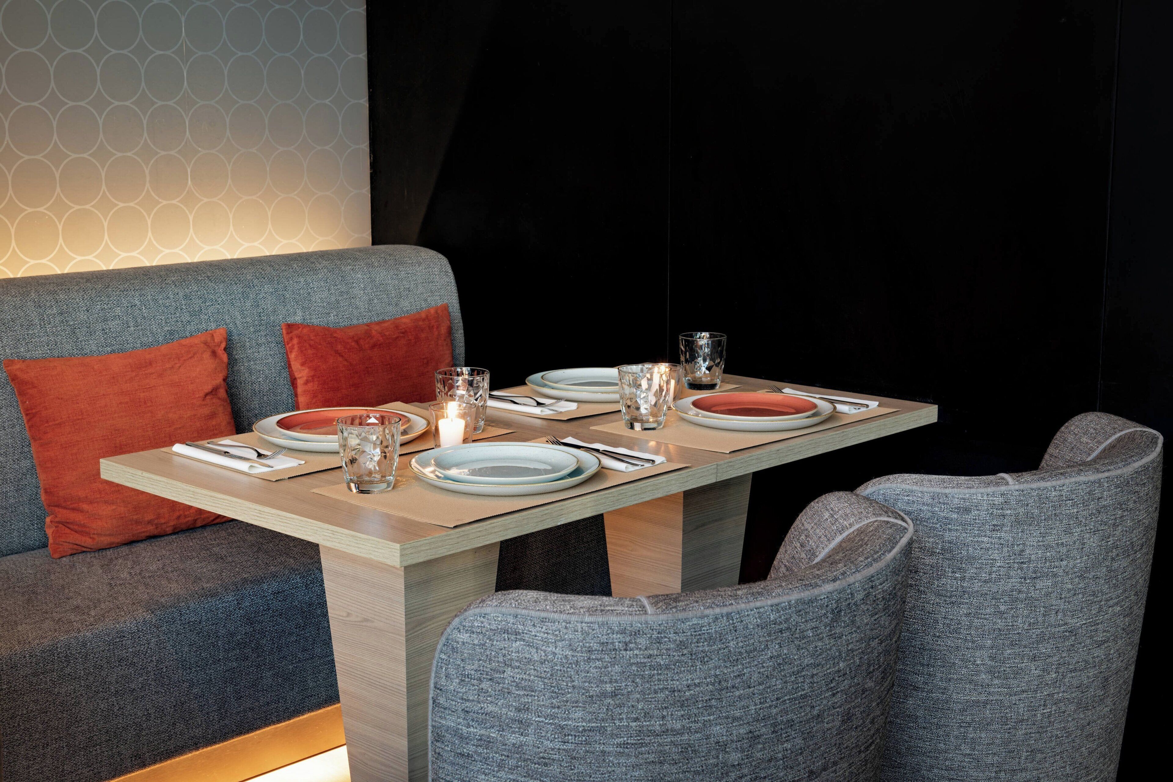 A dining area with a table set for two, a sofa with red cushions, and a patterned wall in the background.