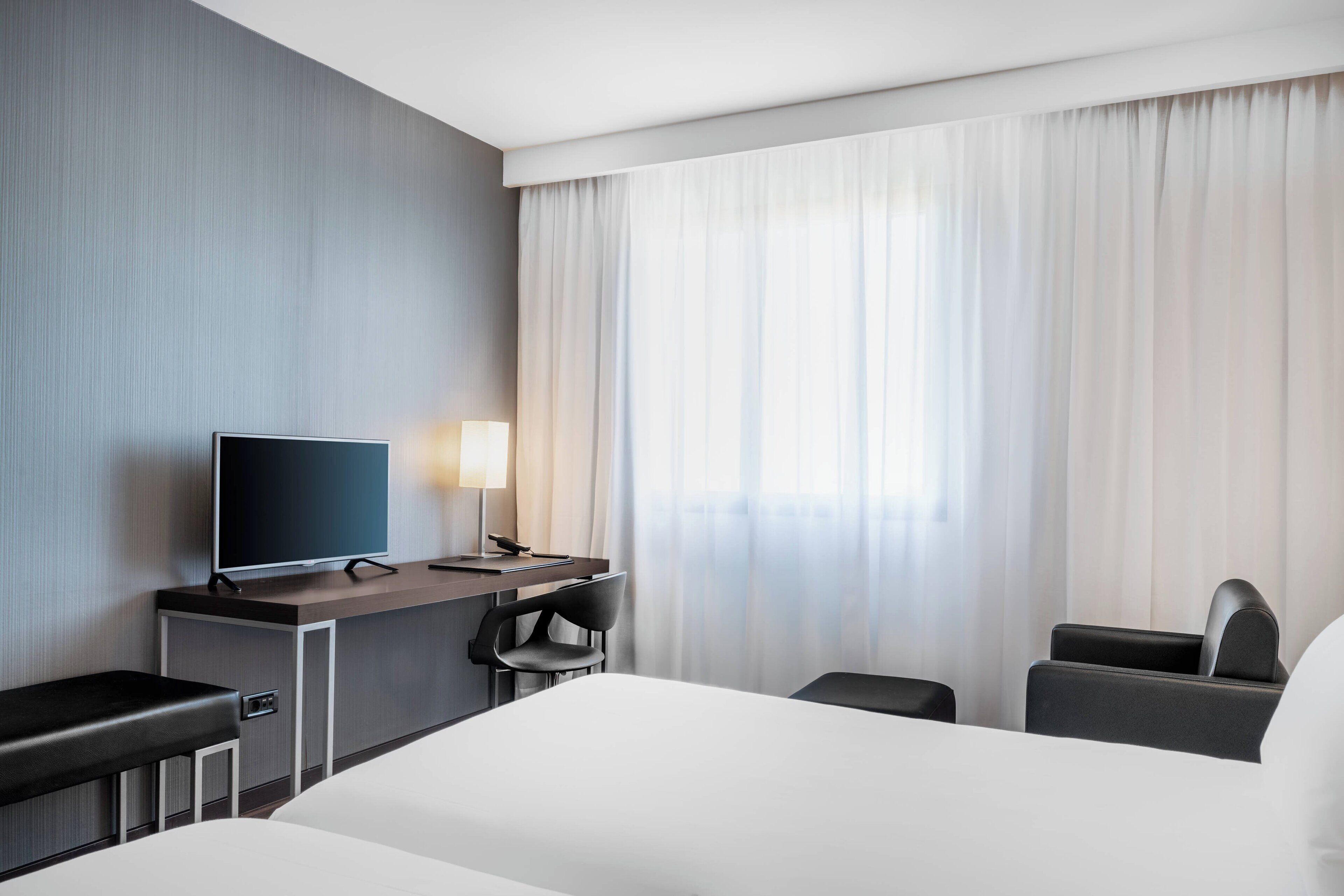 A hotel room with a bed, a desk with a TV, a chair, and a window with curtains.