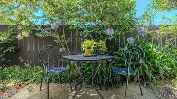 A round metal table with two chairs, surrounded by a garden with various plants and a wooden fence in the background.