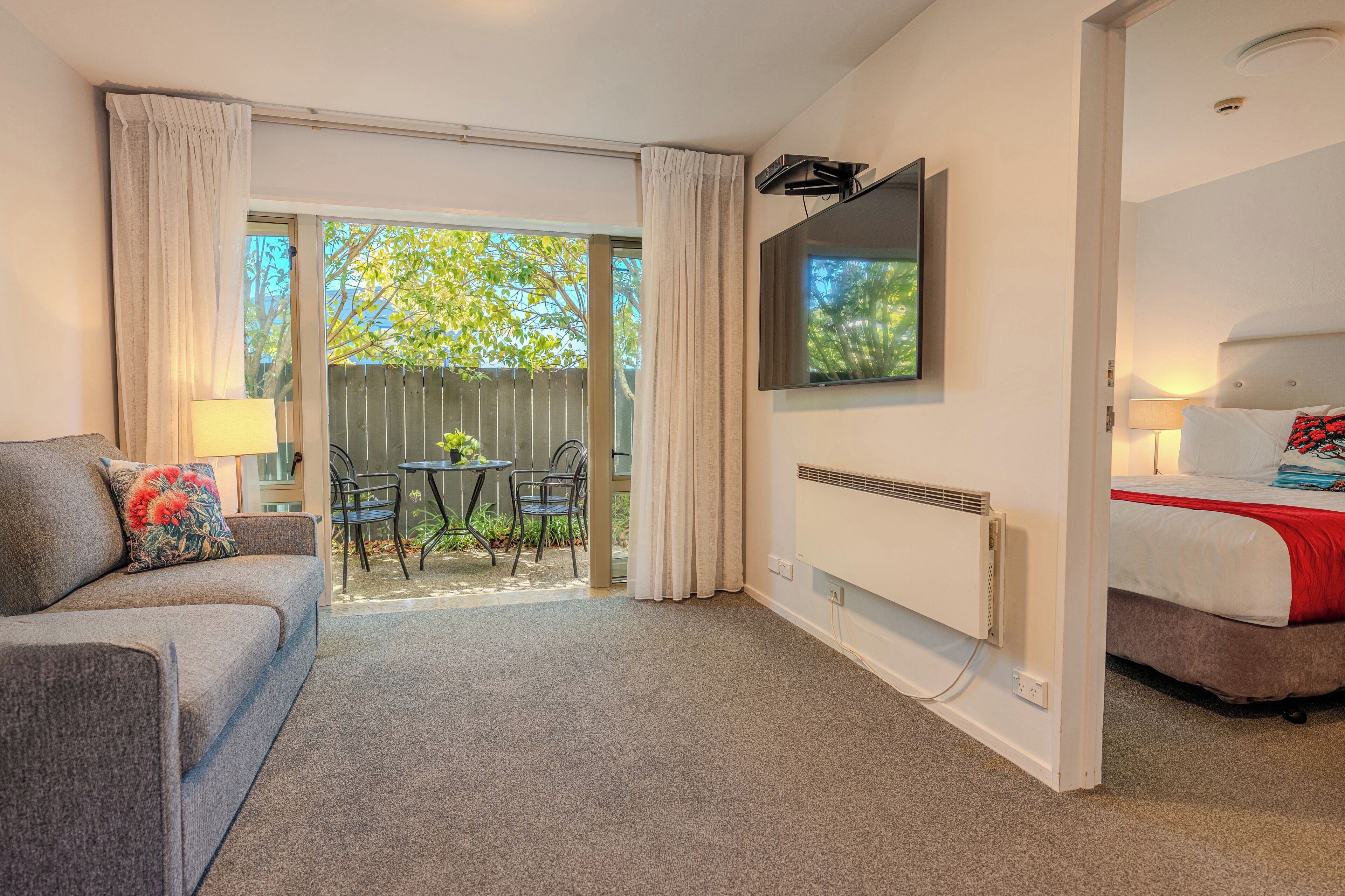 A hotel room with a sofa, a bed, a TV, a radiator, and a sliding glass door leading to an outdoor area with a table and chairs.