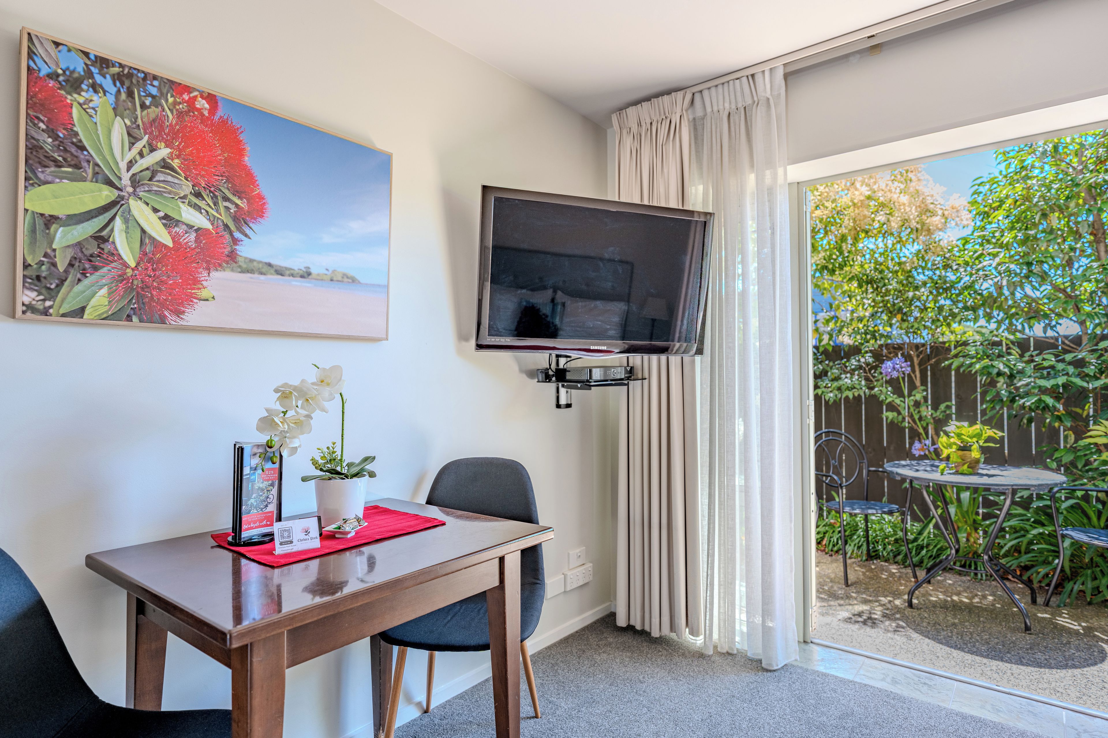 Habitación de hotel con mesa de comedor, sillas, televisor de pantalla plana montado en la pared, una planta en maceta y vista al jardín a través de la puerta corrediza de vidrio.