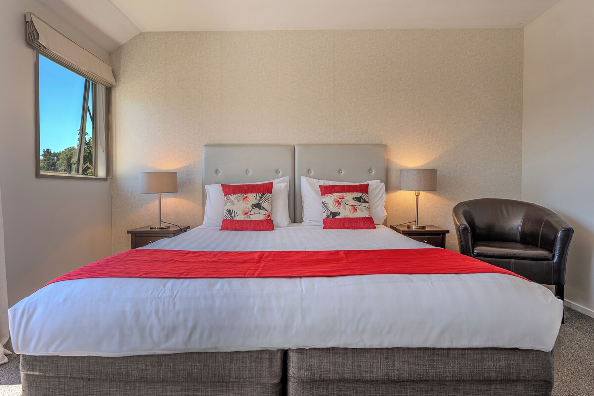 A double bed with a red and white bedspread, two nightstands with lamps, a black leather chair, and a window with a view of trees.