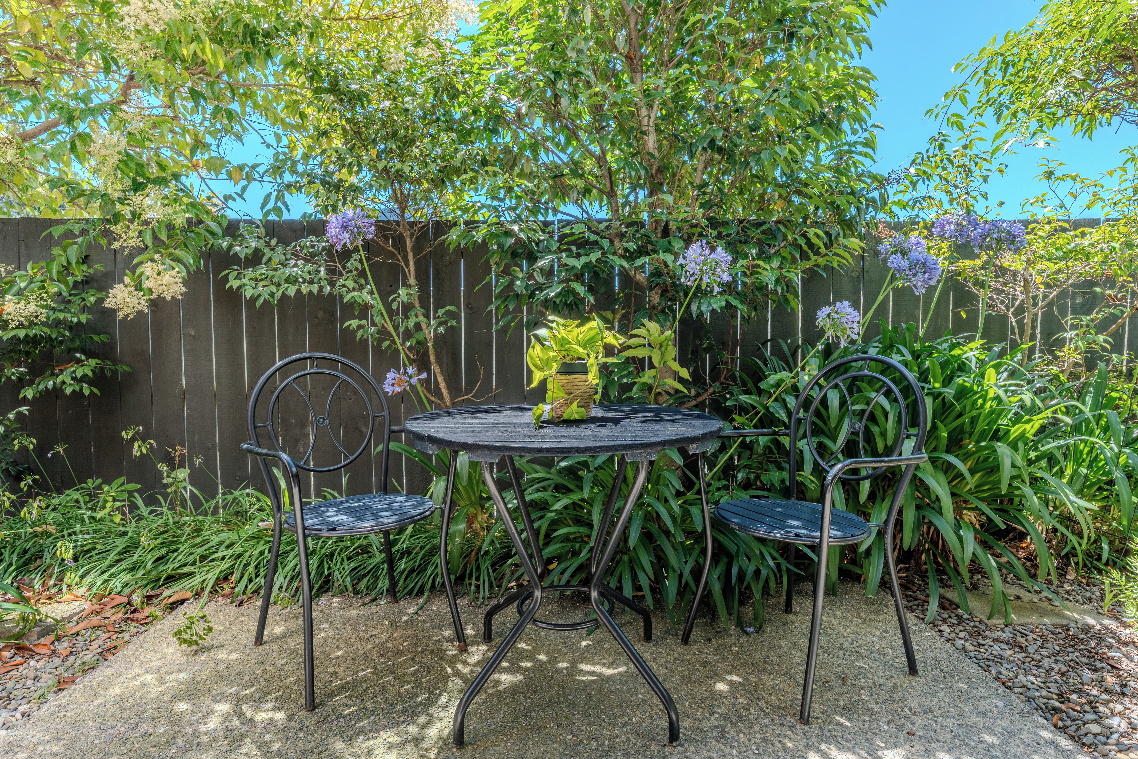 A round metal table with two chairs, surrounded by a garden with various plants and a wooden fence in the background.