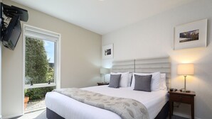 Um quarto bem arrumado com uma cama grande, uma televisão na parede, uma janela com vista para o verde e uma mesa de cabeceira com abajur.