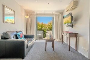 A living room with a sofa, a TV mounted on the wall, a small table, and a balcony with a view. - Chelsea Park Motor Lodge Nelson (Nelson)